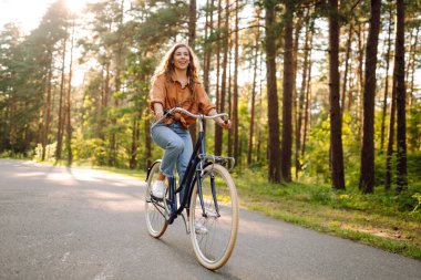 Ormanda, güneşin altında bisiklet süren mutlu bir kadın var. Güzel bir kadın gün batımında bisiklet sürmekten zevk alıyor, dinleniyor. Aktif yaşam tarzı. Hafta sonu konsepti.
