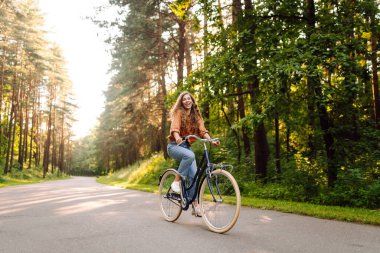 Ormanda, güneşin altında bisiklet süren mutlu bir kadın var. Güzel bir kadın gün batımında bisiklet sürmekten zevk alıyor, dinleniyor. Aktif yaşam tarzı. Hafta sonu konsepti.