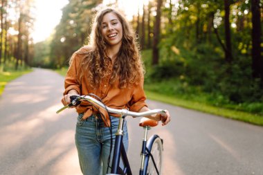 Ormanda, güneşin altında bisiklet süren mutlu bir kadın var. Güzel bir kadın gün batımında bisiklet sürmekten zevk alıyor, dinleniyor. Aktif yaşam tarzı. Hafta sonu konsepti.