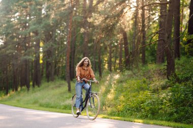 Ormanda, güneşin altında bisiklet süren mutlu bir kadın var. Güzel bir kadın gün batımında bisiklet sürmekten zevk alıyor, dinleniyor. Aktif yaşam tarzı. Hafta sonu konsepti.