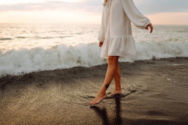 Gün batımında deniz kıyısında yürüyen genç bir kadının bacakları. Deniz kıyısında elbise giymiş güzel bir kadın, doğada özgürlüğün tadını çıkarıyor ve hissediyor. Tatil konsepti, deniz manzarası.