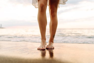 Gün batımında deniz kıyısında yürüyen genç bir kadının bacakları. Deniz kıyısında elbise giymiş güzel bir kadın, doğada özgürlüğün tadını çıkarıyor ve hissediyor. Tatil konsepti, deniz manzarası.