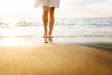Gün batımında deniz kıyısında yürüyen genç bir kadının bacakları. Deniz kıyısında elbise giymiş güzel bir kadın, doğada özgürlüğün tadını çıkarıyor ve hissediyor. Tatil konsepti, deniz manzarası.
