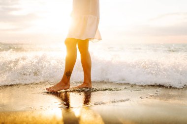 Gün batımında deniz kıyısında yürüyen genç bir kadının bacakları. Deniz kıyısında elbise giymiş güzel bir kadın, doğada özgürlüğün tadını çıkarıyor ve hissediyor. Tatil konsepti, deniz manzarası.