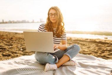 Gün batımında elinde dizüstü bilgisayarla sahilde oturan bir kadının portresi. Dizüstü bilgisayarda çalışan, klavyede dışarıda oturan güzel bir bayan serbest yazar. Bağımsız kavram, doğa ve rahatlama.