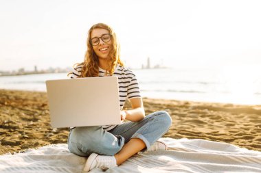 Gün batımında elinde dizüstü bilgisayarla sahilde oturan bir kadının portresi. Dizüstü bilgisayarda çalışan, klavyede dışarıda oturan güzel bir bayan serbest yazar. Bağımsız kavram, doğa ve rahatlama.