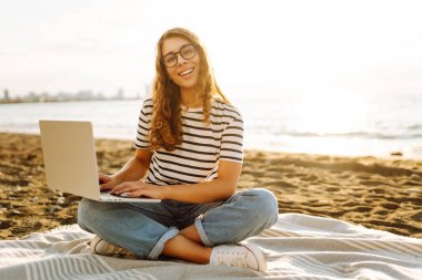 Gün batımında elinde dizüstü bilgisayarla sahilde oturan bir kadının portresi. Dizüstü bilgisayarda çalışan, klavyede dışarıda oturan güzel bir bayan serbest yazar. Bağımsız kavram, doğa ve rahatlama.