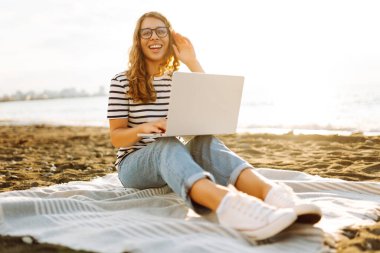 Gün batımında elinde dizüstü bilgisayarla sahilde oturan bir kadının portresi. Dizüstü bilgisayarda çalışan, klavyede dışarıda oturan güzel bir bayan serbest yazar. Bağımsız kavram, doğa ve rahatlama.