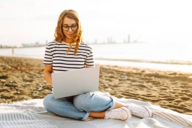 Gün batımında elinde dizüstü bilgisayarla sahilde oturan bir kadının portresi. Dizüstü bilgisayarda çalışan, klavyede dışarıda oturan güzel bir bayan serbest yazar. Bağımsız kavram, doğa ve rahatlama.