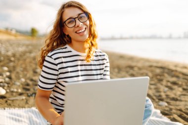 Gün batımında elinde dizüstü bilgisayarla sahilde oturan bir kadının portresi. Dizüstü bilgisayarda çalışan, klavyede dışarıda oturan güzel bir bayan serbest yazar. Bağımsız kavram, doğa ve rahatlama.