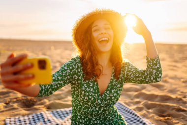 Gün batımı plajında elinde telefonla selfie çeken genç bir kadın. Parlak elbiseli ve şapkalı neşeli bir kadın deniz manzarasının tadını çıkarıyor ve telefonda sohbet ediyor. Bloglama ve boş zaman kavramı.