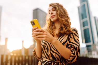 Bir metropolün arka planına telefon tutan genç bir kadının ellerinin yakın çekimi. Güzel bir kadın telefonda mesaj atıyor ve dışarıda gezmekten zevk alıyor. Teknoloji kavramı, şehirler.