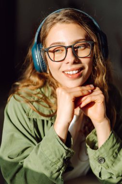 Gözlüklü genç bir kadın kablosuz kulaklıkla müzik dinliyor ve sıcak bir dairede güneşin tadını çıkarıyor. Gözlüklü gülümseyen kadın oturma odasında kendi kendine müzik dinleyerek vakit geçiriyor. Yaşam biçimi.