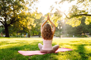 Nilüfer çiçekli poz veren genç bir kadın ellerini parlak yoga minderinin üstüne kaldırmış. Sakin kadın yoga yapıyor ve güneşli parkta egzersiz yapıyor. Rahatlama, spor ve doğa konsepti.