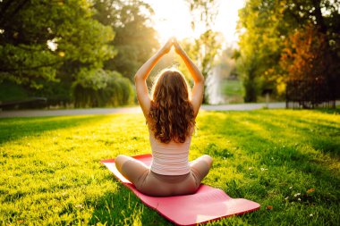 Nilüfer çiçekli poz veren genç bir kadın ellerini parlak yoga minderinin üstüne kaldırmış. Sakin kadın yoga yapıyor ve güneşli parkta egzersiz yapıyor. Rahatlama, spor ve doğa konsepti.