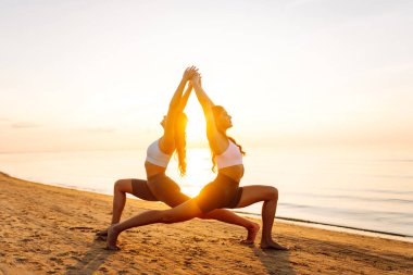 İki atletik kadın deniz kıyısında birlikte yoga yapıyorlar. Güzel arkadaşlar gün batımında spor meditasyonu yapıyorlar. Spor, zevk ve meditasyon kavramı.