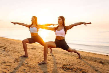 İki atletik kadın deniz kıyısında birlikte yoga yapıyorlar. Güzel arkadaşlar gün batımında spor meditasyonu yapıyorlar. Spor, zevk ve meditasyon kavramı.