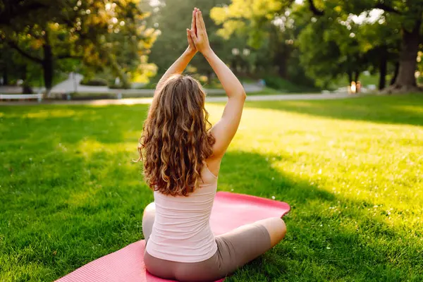 Nilüfer çiçekli poz veren genç bir kadın ellerini parlak yoga minderinin üstüne kaldırmış. Sakin kadın yoga yapıyor ve güneşli parkta egzersiz yapıyor. Rahatlama, spor ve doğa konsepti.