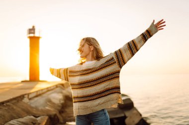 İskelede durup günbatımının tadını çıkaran rahat bir kadın. Genç kadın kendini özgür hissediyor ve deniz fenerinin deniz manzarasına bakıyor. Özgürlük kavramı, keyif.