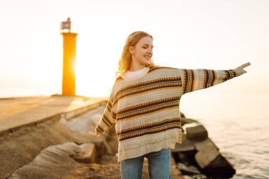 İskelede durup günbatımının tadını çıkaran rahat bir kadın. Genç kadın kendini özgür hissediyor ve deniz fenerinin deniz manzarasına bakıyor. Özgürlük kavramı, keyif.