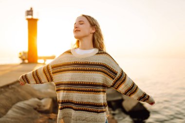 İskelede durup günbatımının tadını çıkaran rahat bir kadın. Genç kadın kendini özgür hissediyor ve deniz fenerinin deniz manzarasına bakıyor. Özgürlük kavramı, keyif.