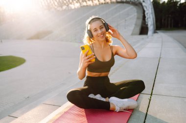 Kulaklık takan ve hasırla egzersiz yapan bir kadının portresi. Spor üniformalı genç bir kadın pembe yoga minderine oturur ve müzik dinler..