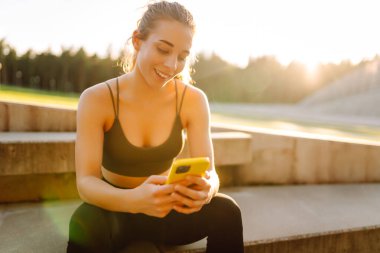 Kulaklık takan ve hasırla egzersiz yapan bir kadının portresi. Spor üniformalı genç bir kadın pembe yoga minderine oturur ve müzik dinler..