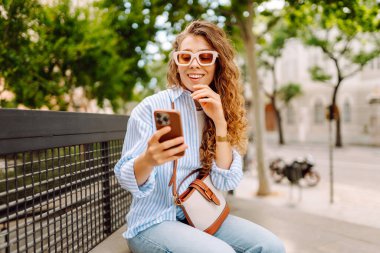 Elinde telefonla şehirde yürüyüşün tadını çıkaran genç bir kadının portresi. Neşeli kadın bankta oturuyor ve telefonda mesajlaşıyor. Yaşam biçimi.