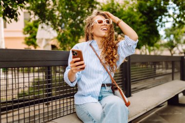 Elinde telefonla şehirde yürüyüşün tadını çıkaran genç bir kadının portresi. Neşeli kadın bankta oturuyor ve telefonda mesajlaşıyor. Yaşam biçimi.