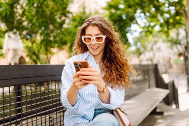 Elinde telefonla şehirde yürüyüşün tadını çıkaran genç bir kadının portresi. Neşeli kadın bankta oturuyor ve telefonda mesajlaşıyor. Yaşam biçimi.