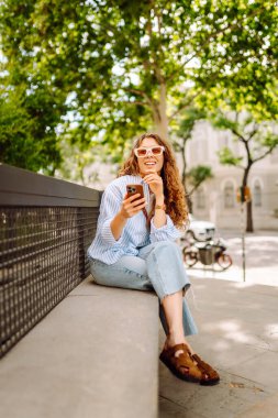 Elinde telefonla şehirde yürüyüşün tadını çıkaran genç bir kadının portresi. Neşeli kadın bankta oturuyor ve telefonda mesajlaşıyor. Yaşam biçimi.