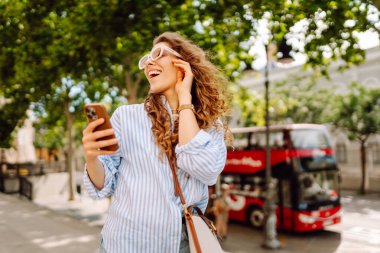 Elinde telefonla şehirde yürüyüşün tadını çıkaran genç bir kadının portresi. Neşeli kadın bankta oturuyor ve telefonda mesajlaşıyor. Yaşam biçimi.