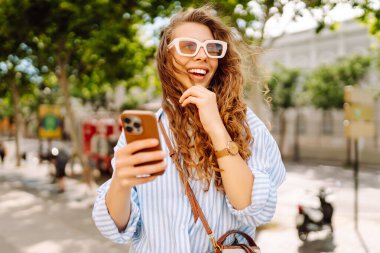 Elinde telefonla şehirde yürüyüşün tadını çıkaran genç bir kadının portresi. Neşeli kadın bankta oturuyor ve telefonda mesajlaşıyor. Yaşam biçimi.