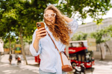 Elinde telefonla şehirde yürüyüşün tadını çıkaran genç bir kadının portresi. Neşeli kadın bankta oturuyor ve telefonda mesajlaşıyor. Yaşam biçimi.