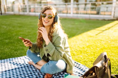 Günlük giysiler içinde, kablosuz kulaklık takan, telefon kullanan çimenlikteki bir paspasın üzerinde oturan palmiye ağaçlarıyla çevrili genç bir kadın. Güzel bir kadın müzikten ve doğada zamandan zevk alıyor..