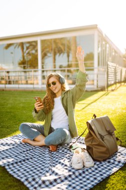 Günlük giysiler içinde, kablosuz kulaklık takan, telefon kullanan çimenlikteki bir paspasın üzerinde oturan palmiye ağaçlarıyla çevrili genç bir kadın. Güzel bir kadın müzikten ve doğada zamandan zevk alıyor..