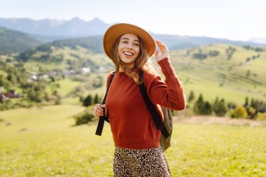 Şapkalı ve sırt çantalı genç bir kadın yamaçta güneşli havanın tadını çıkarıyor. Güzel bayan yürüyüşçü gün batımında dağ yolunda özgürlüğünü hissediyor. Rahatlama ve doğa kavramı.