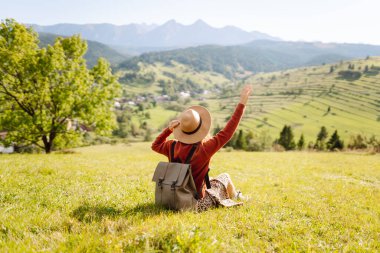 Şapkalı ve sırt çantalı genç bir kadın yamaçta güneşli havanın tadını çıkarıyor. Güzel bayan yürüyüşçü gün batımında dağ yolunda özgürlüğünü hissediyor. Rahatlama ve doğa kavramı.