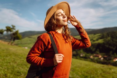 Şapkalı ve sırt çantalı genç bir kadın yamaçta güneşli havanın tadını çıkarıyor. Güzel bayan yürüyüşçü gün batımında dağ yolunda özgürlüğünü hissediyor. Rahatlama ve doğa kavramı.
