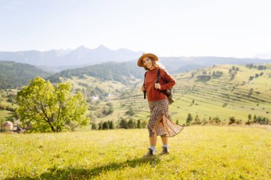 Şapkalı ve sırt çantalı genç bir kadın yamaçta güneşli havanın tadını çıkarıyor. Güzel bayan yürüyüşçü gün batımında dağ yolunda özgürlüğünü hissediyor. Rahatlama ve doğa kavramı.
