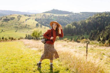 Şapkalı ve sırt çantalı genç bir kadın yamaçta güneşli havanın tadını çıkarıyor. Güzel bayan yürüyüşçü gün batımında dağ yolunda özgürlüğünü hissediyor. Rahatlama ve doğa kavramı.