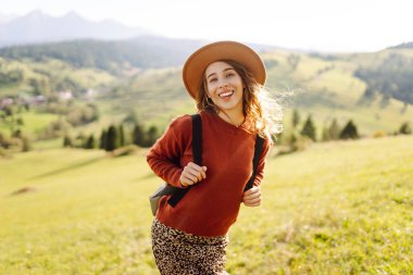 Şapkalı ve sırt çantalı genç bir kadın yamaçta güneşli havanın tadını çıkarıyor. Güzel bayan yürüyüşçü gün batımında dağ yolunda özgürlüğünü hissediyor. Rahatlama ve doğa kavramı.