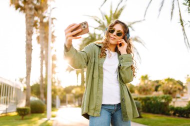 Güneş gözlüklü ve kablosuz kulaklıklı mutlu kadın elinde telefonla palmiye ağaçlarıyla caddede yürüyor. Güzel gezgin dışarıda müzik dinler ve sohbet eder. Etkin yaşam biçimi.