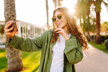 Güneş gözlüklü ve kablosuz kulaklıklı mutlu kadın elinde telefonla palmiye ağaçlarıyla caddede yürüyor. Güzel gezgin dışarıda müzik dinler ve sohbet eder. Etkin yaşam biçimi.