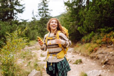 Sırtında parlak sarı bir sırt çantasıyla yürüyüş parkurunda telefonla gülümseyen kadın yürüyüşçü. Macerayı seven ve dağlarda yürüyüş yapan güzel bir kadın. Seyahat ve günlük tutma kavramı.