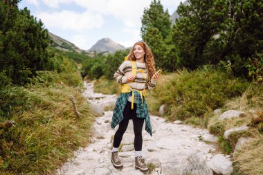 Sırtında parlak sarı bir sırt çantasıyla yürüyüş parkurunda telefonla gülümseyen kadın yürüyüşçü. Macerayı seven ve dağlarda yürüyüş yapan güzel bir kadın. Seyahat ve günlük tutma kavramı.