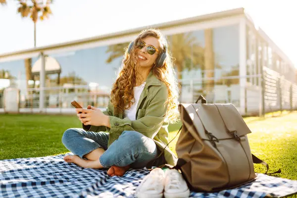 Günlük giysiler içinde, kablosuz kulaklık takan, telefon kullanan çimenlikteki bir paspasın üzerinde oturan palmiye ağaçlarıyla çevrili genç bir kadın. Güzel bir kadın müzikten ve doğada zamandan zevk alıyor..