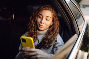 Telefonlu genç bir kadın arabanın arka koltuğunda oturmuş pencereden dışarı bakıyor. Taksideki mutlu kadın telefonda mesaj atıyor. Ulaşım, ulaşım, sohbet kavramı.