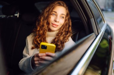 Telefonlu genç bir kadın arabanın arka koltuğunda oturmuş pencereden dışarı bakıyor. Taksideki mutlu kadın telefonda mesaj atıyor. Ulaşım, ulaşım, sohbet kavramı.