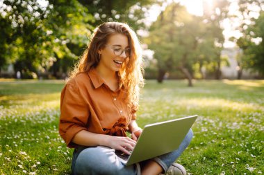 Parktaki çimlerin üzerinde rahatlayan dizüstü bilgisayarı olan hoş bir kadının portresi. Gülümseyen serbest çalışan kadın dışarıda çalışmaktan zevk alıyor. Uzak çalışma ve doğa kavramı.
